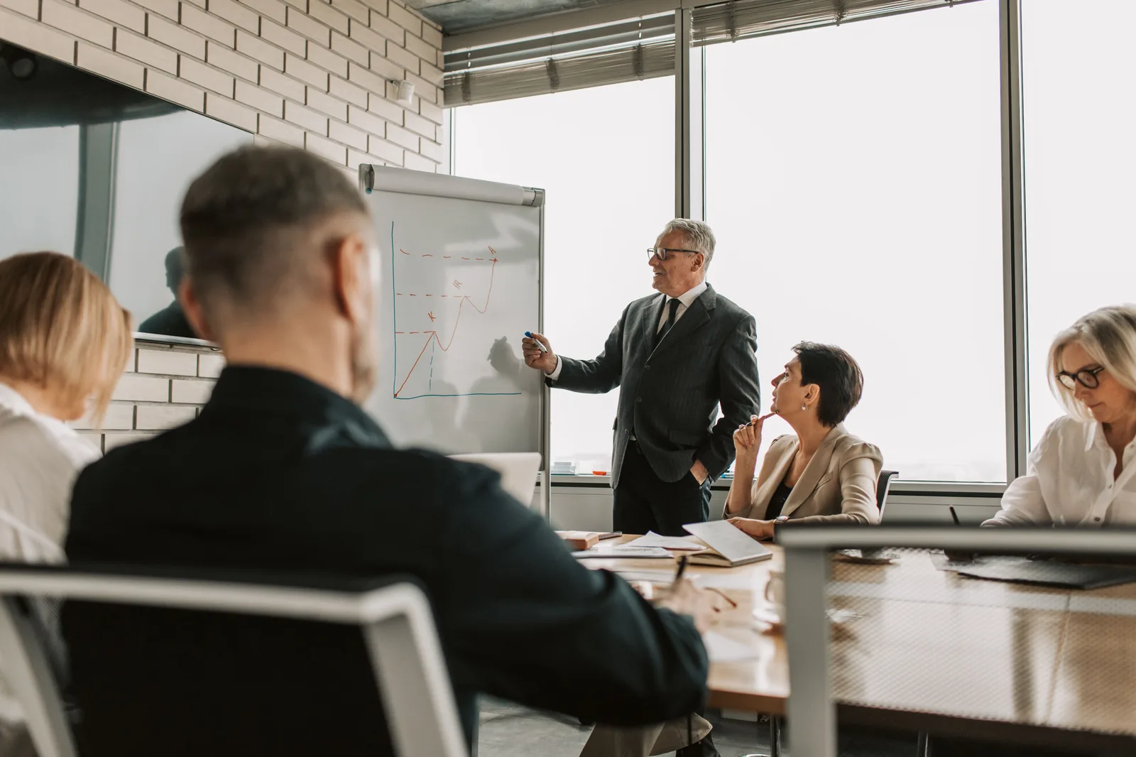 Executive coach standing at a whiteboard teaching a strategy session to leaders seated at a conference table